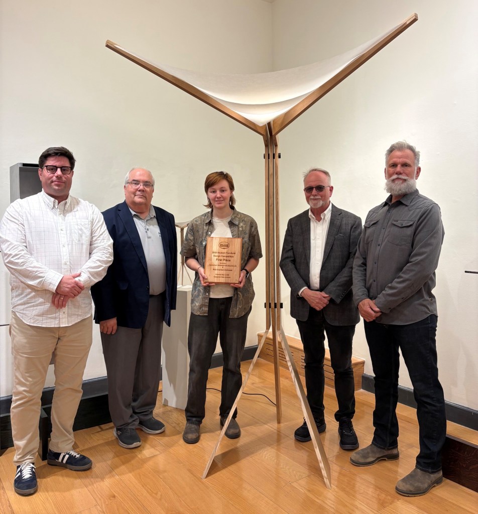 A group of five people standing together, with one person holding a wooden award. The backdrop features an art installation with a unique structure made of wood and fabric.