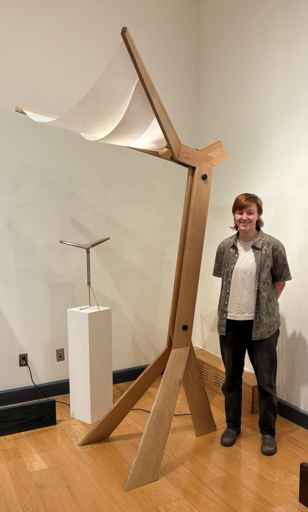A young person stands next to a large wooden sculpture that features a curved paper element, in a gallery setting. Another smaller sculpture is visible in the background, alongside wooden flooring.