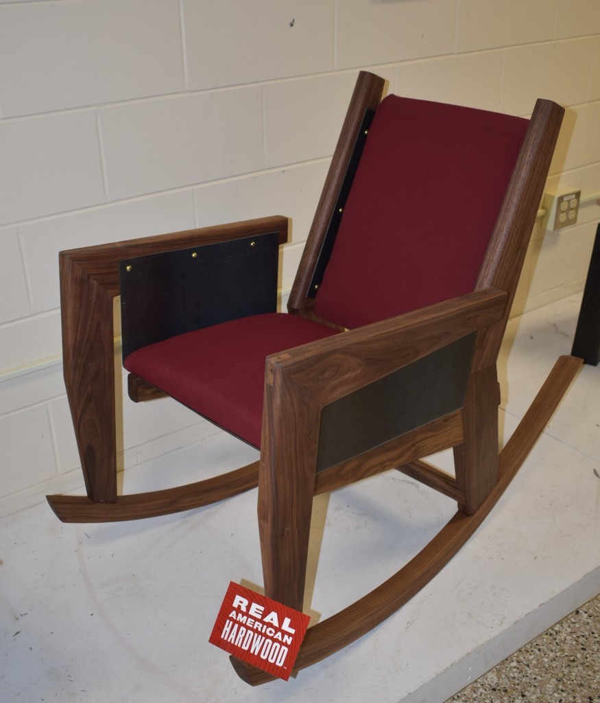 A wooden rocking chair with a burgundy cushioned seat and backrest, featuring a sign that reads 'REAL AMERICAN HARDWOOD'.