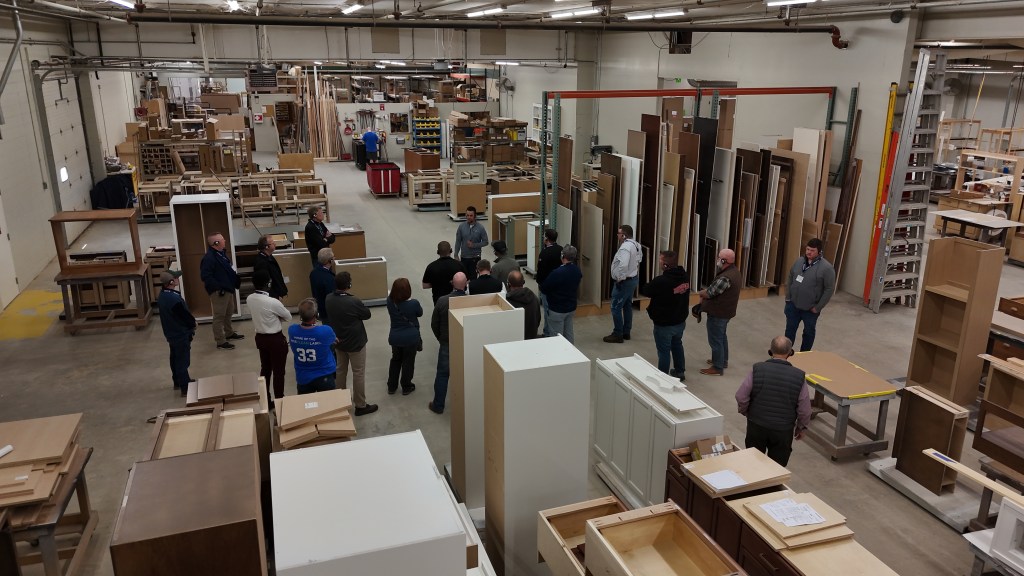 A group of people gathered in a large workshop filled with wood and cabinet materials, engaging in a discussion while surrounded by various unfinished products.