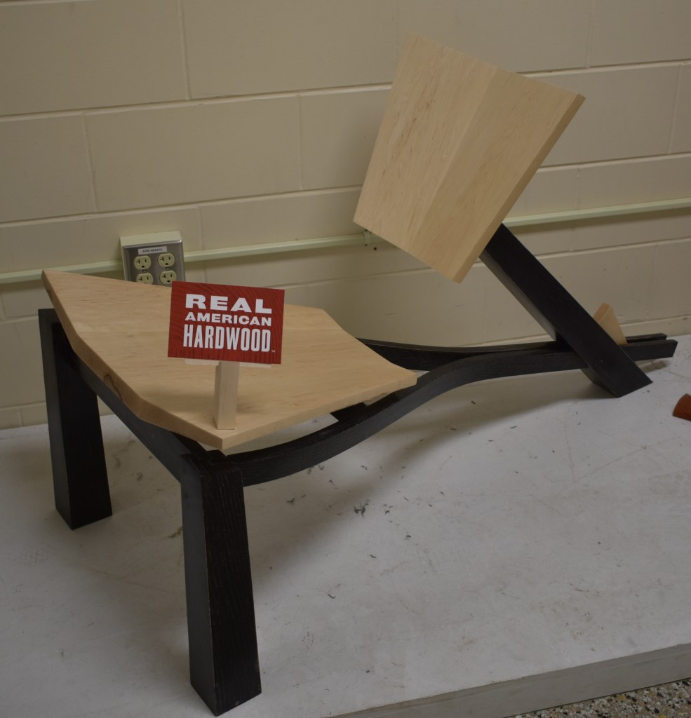 A modern wooden chair made of light-colored hardwood with a slanted backrest, positioned on a tabletop with a sign that reads 'REAL AMERICAN HARDWOOD'. The chair has a unique design featuring curved black legs.