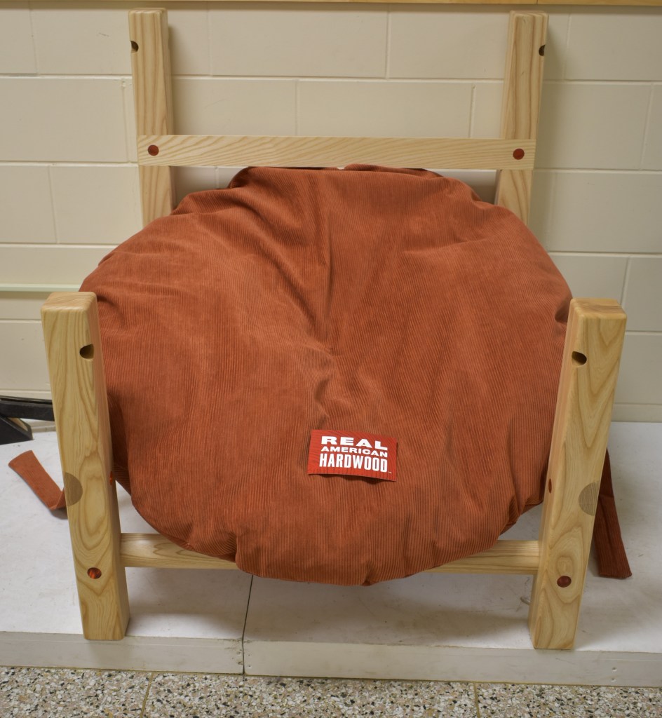 A wooden chair frame with an orange corduroy cushion, featuring a label that reads 'REAL AMERICAN HARDWOOD'.