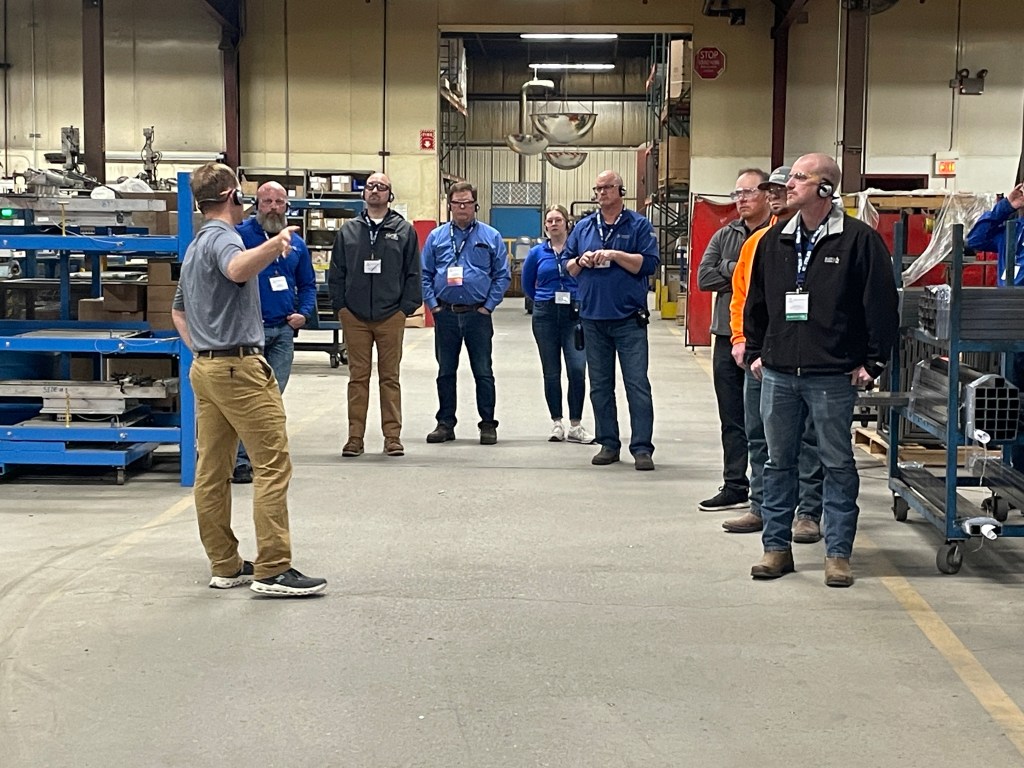 A group of people listening to a speaker in a manufacturing facility, with various equipment and storage racks visible in the background.